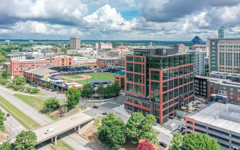 Northwood Ravin Property Management Portfolio, 555 MANGUM in Durham, NC, a multi-family, multi-story apartment showing the building exterior with plentiful windows, retail shops on the first floor, and a large baseball stadium across the street