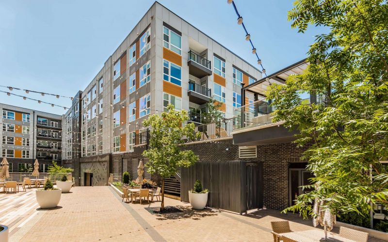 The Northwood Ravin apartment complex boasts a modern design with a communal courtyard featuring seating areas, lush greenery, and enchanting hanging string lights. Four-story buildings surround this inviting space.