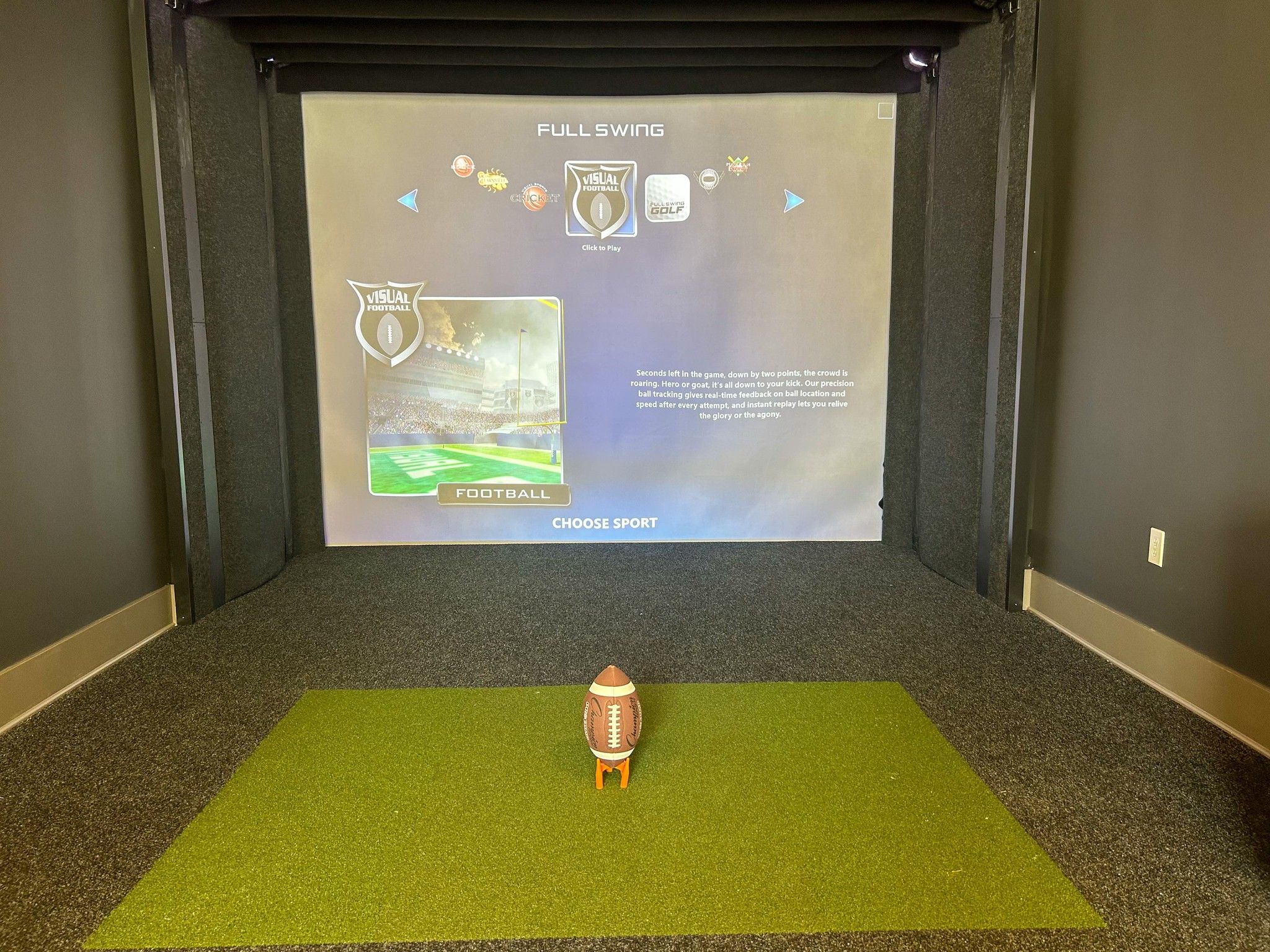 A Northwood Ravin football on a stand is placed on a green mat in front of a large projection screen, displaying a sports game menu with options to select different sports.