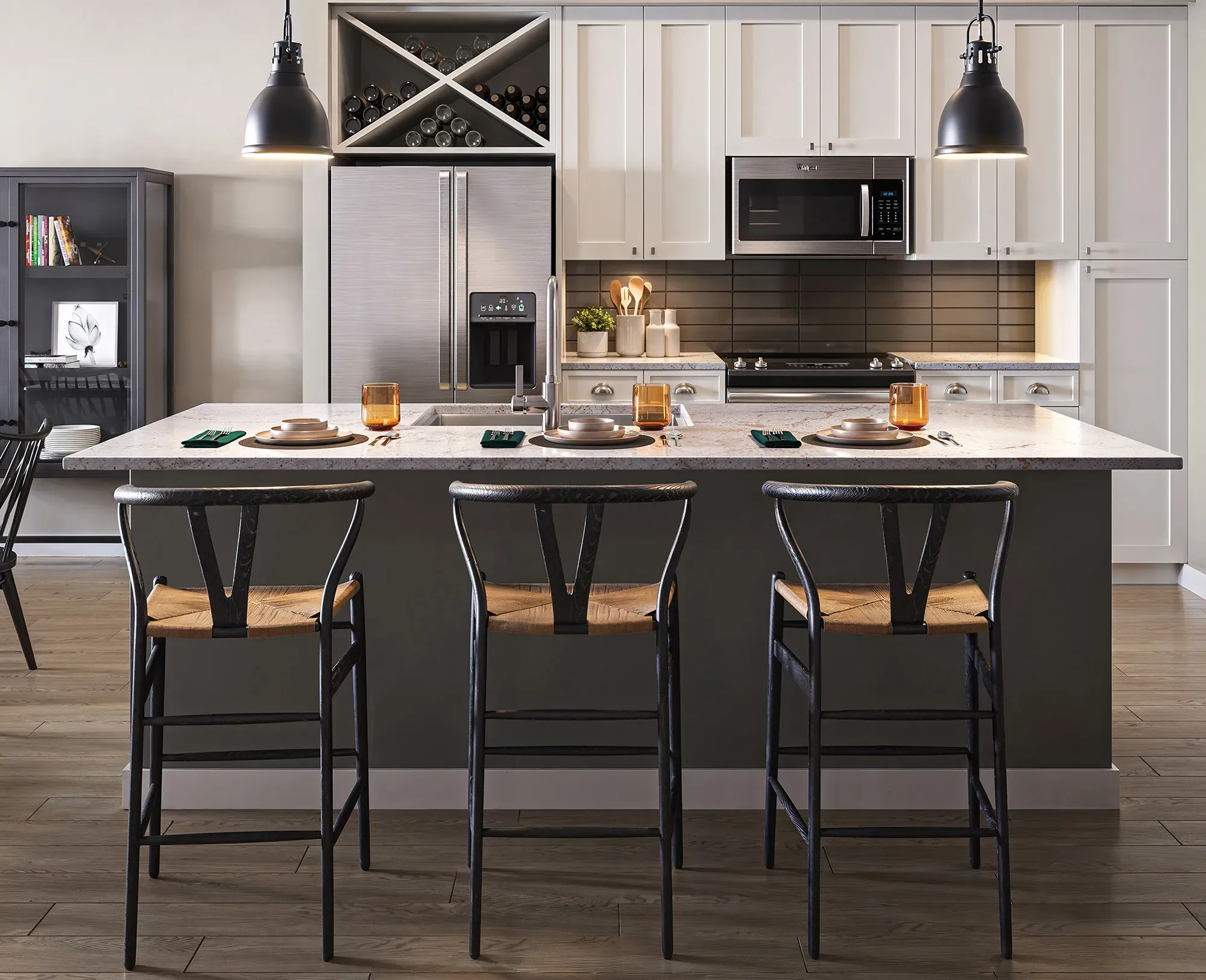 Modern kitchen with a large island featuring seating accommodations, white cabinetry, stainless steel appliances, overhead lighting, and a small bookcase in the background.