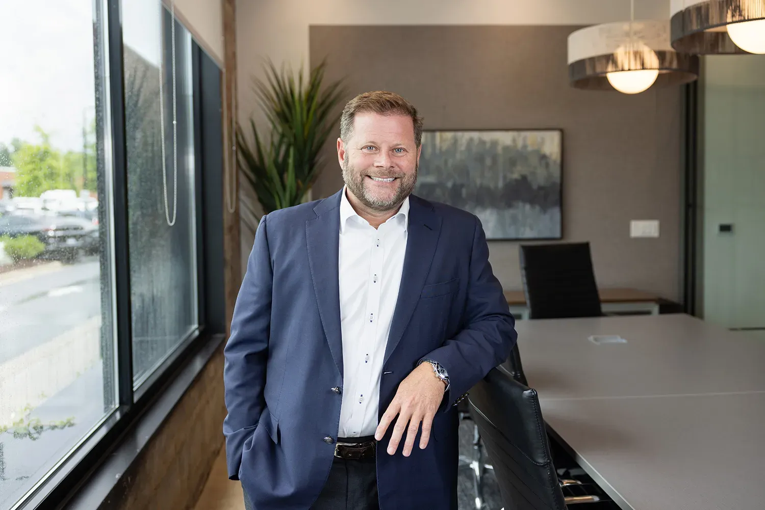 David Ravin in a blazer and white shirt stands smiling in a modern office with large windows, a table, chairs, and decorative lighting.