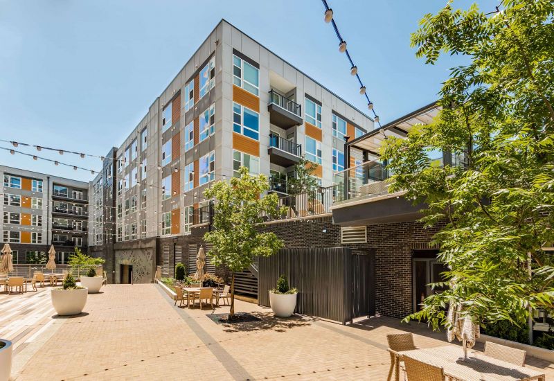 The Northwood Ravin apartment complex boasts a modern design with a communal courtyard featuring seating areas, lush greenery, and enchanting hanging string lights. Four-story buildings surround this inviting space.