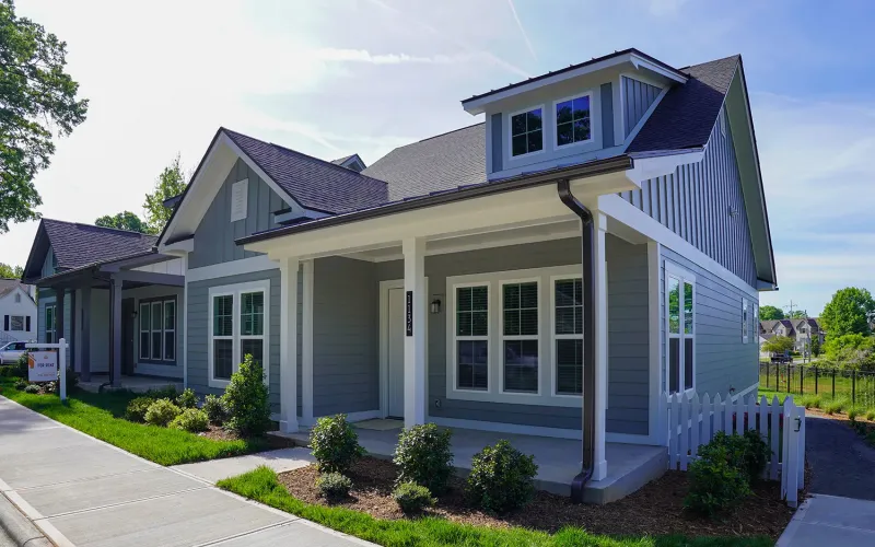 A modern, two-story, gray house with white trim, a covered front porch, and landscaped yard on a sunny day.