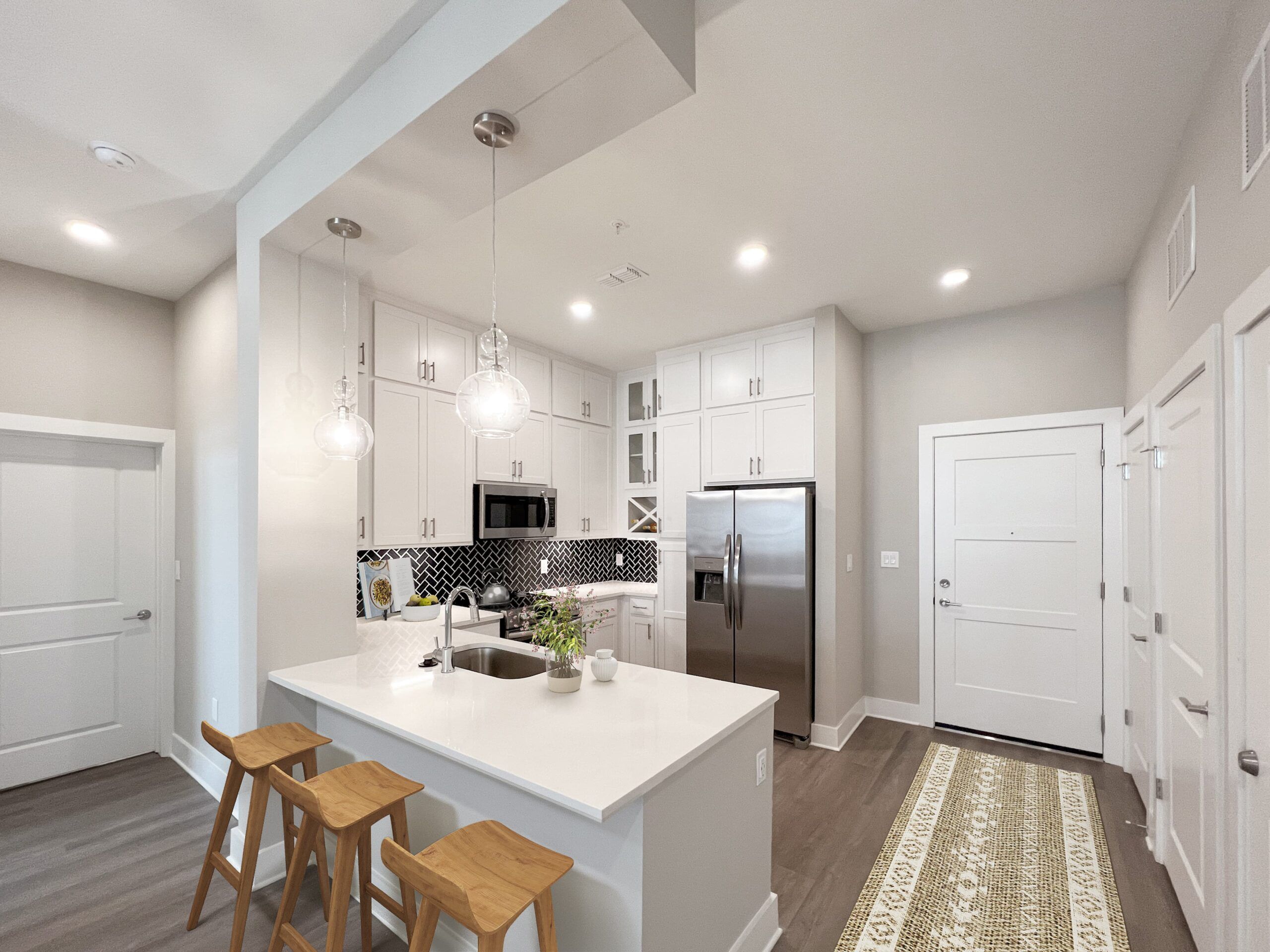 Modern Northwood Ravin kitchen with white cabinetry, stainless steel appliances, a breakfast bar with three wooden stools, pendant lighting, and a patterned rug near the doorway.