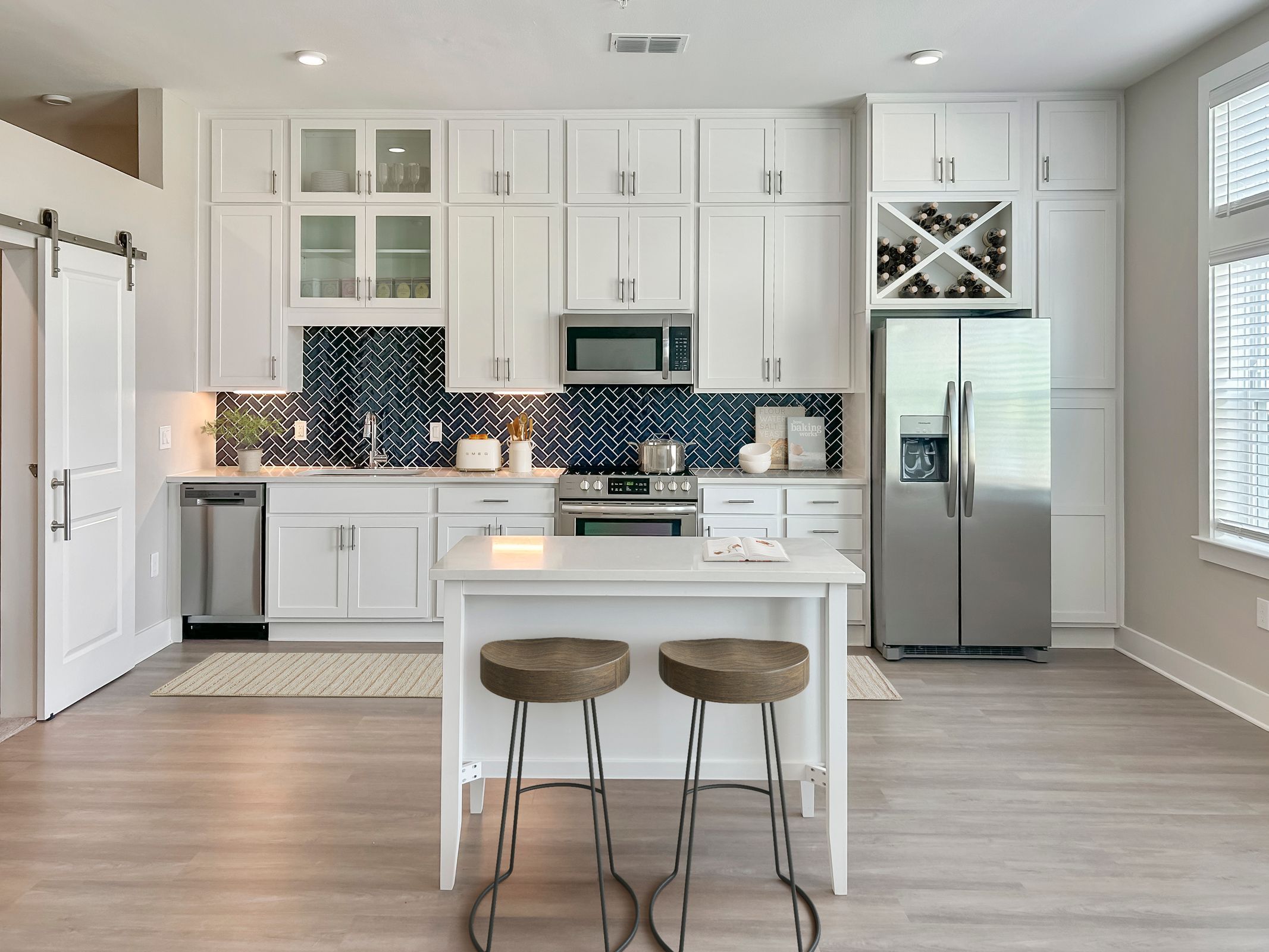 A Northwood Ravin modern kitchen with white cabinets, stainless steel appliances, a herringbone tile backsplash, a small island with two wooden stools, and a wine rack above the refrigerator.