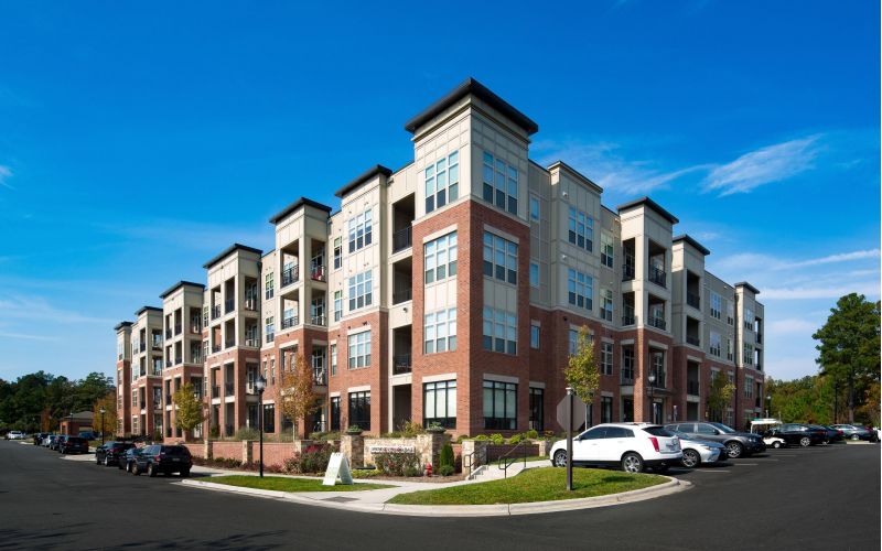 A four-story modern apartment building with brick and beige exterior stands proudly under a clear blue sky in the Northwood Ravin community, surrounded by parked cars and a well-maintained landscape.