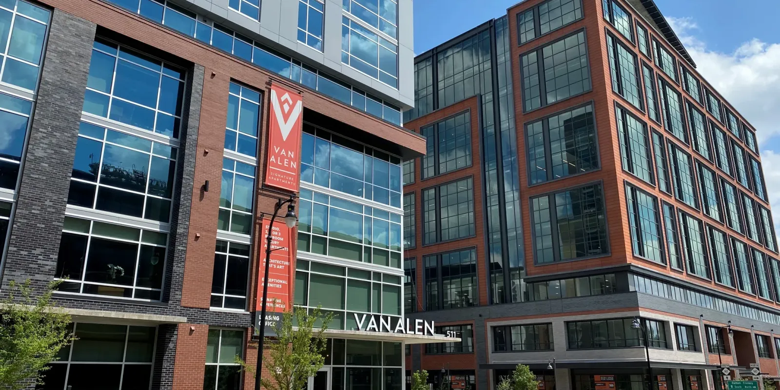 Modern multi-story buildings with large windows. A red banner on the left building reads "VAN ALEN." The street in front is empty with a few trees and a bench visible.