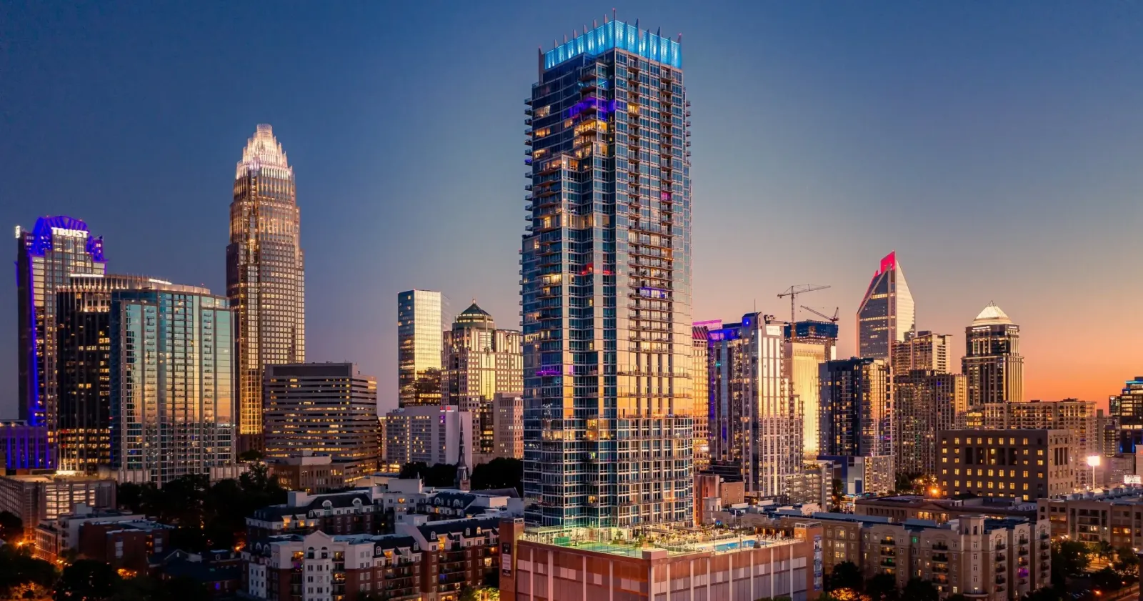 A cityscape at dusk featuring illuminated skyscrapers, with the tallest building in the center displaying vibrant blue, purple, and yellow lighting accents against a clear twilight sky—a vision reminiscent of Northwood Ravin's signature style.