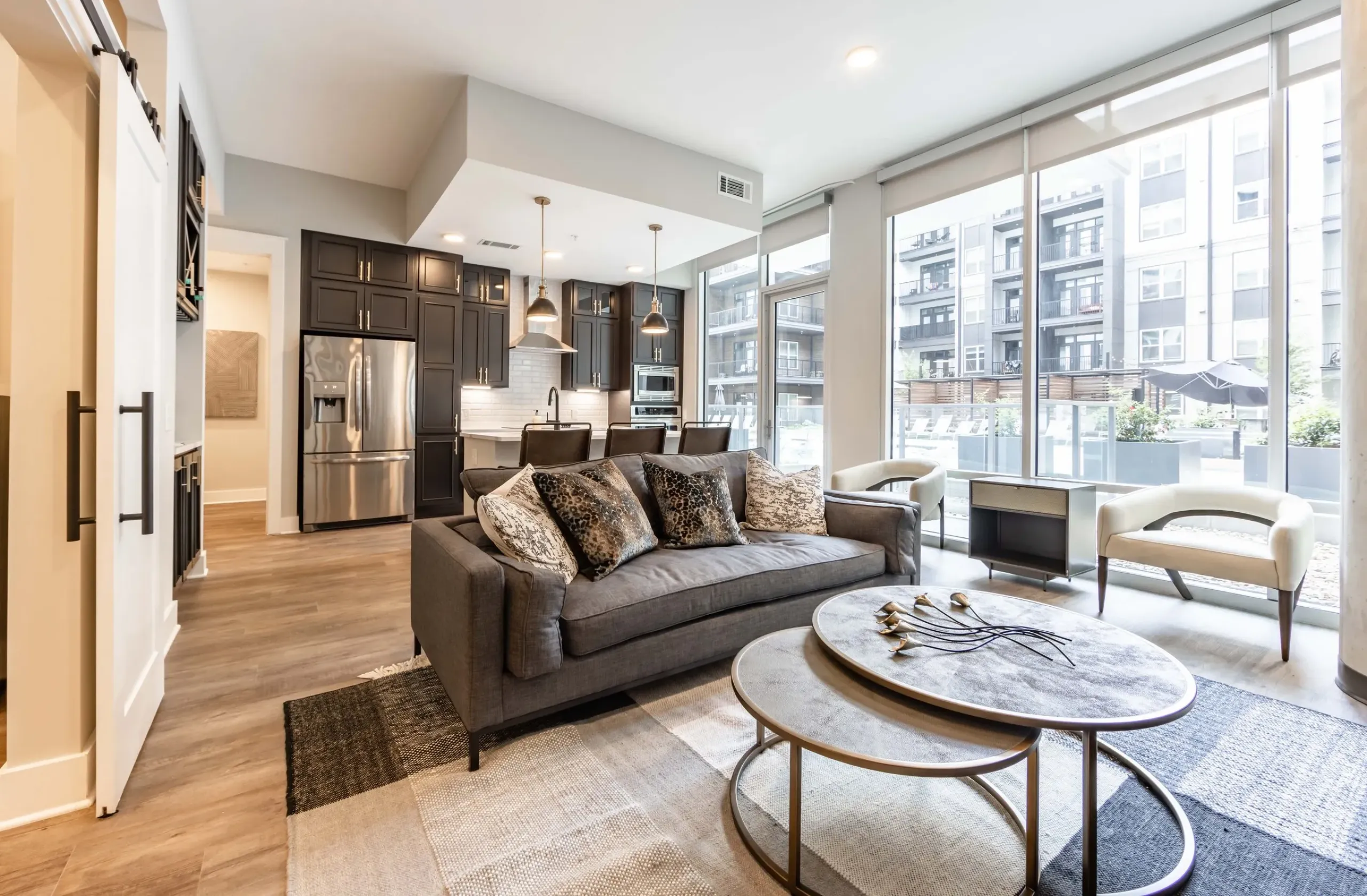 Modern living room with a gray sofa, round coffee tables, and floor-to-ceiling windows overlooking the Northwood Ravin complex. The open kitchen features dark cabinets, a stainless steel refrigerator, and pendant lights.