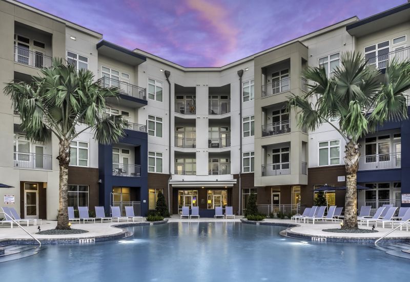 Northwood Ravin presents a modern apartment complex featuring a central swimming pool, surrounded by lounge chairs and palm trees, all under a purple-and-pink sunset sky.