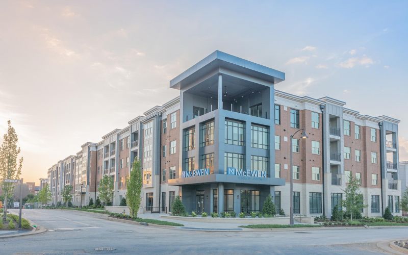 A modern, multi-story apartment building with the name "McEwen" prominently displayed in blue letters by Northwood Ravin. The building features large windows, a central entrance, and a landscaped exterior.